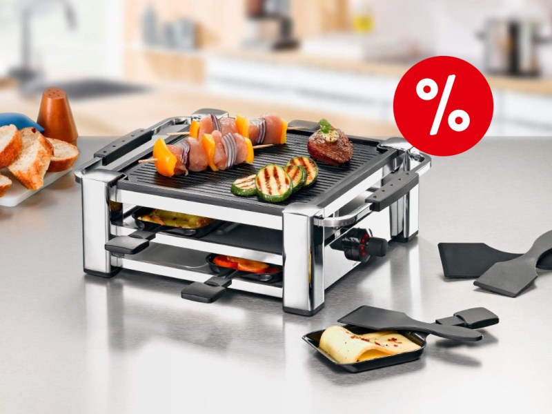 Silbern schwarzer Raclette-Grill schräg von vorne mit zwei Etagen mit befüllten Pfännchen und Gemüse oben auf einer Grillplatte auf hellem spiegelndem Tisch umringt von gebackenenm Käse in Pfännchen und Spateln