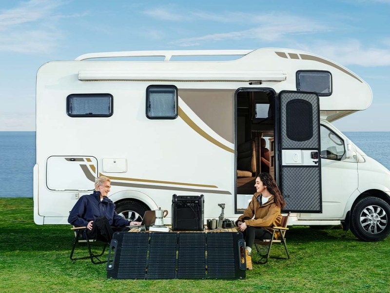 Campervan auf einer Wiese stehend, davor sitzen zwei Personen an einem Tisch, vor ihnen steht ein Solarpanel und eine Powerstation