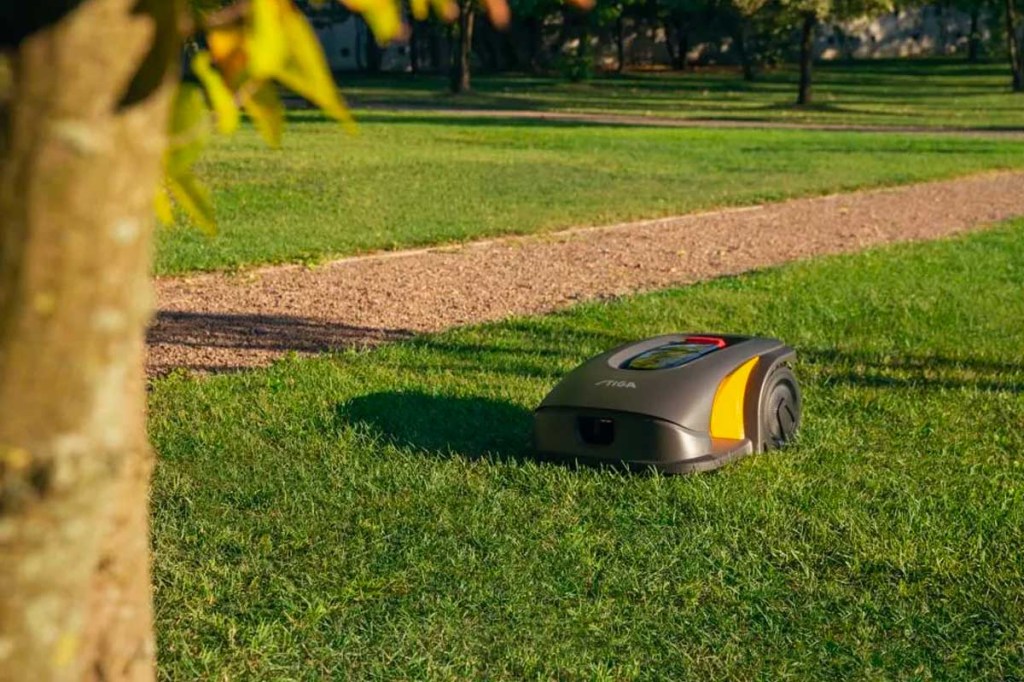 Der Stiga Expert A 5000 fährt auf einem Rasenstück neben einem Sandweg. Im Vordergrund ist ein Baum zu sehen.