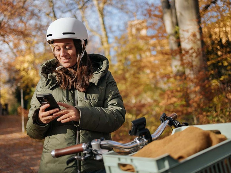 Frau steht in einem herbstlichen Wald neben ihrem Fahrrad und schaut aufs Handy