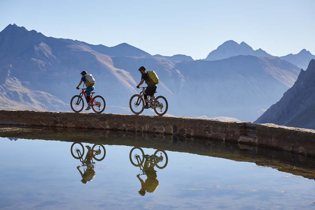Zwei Radfahrer fahren an einem See entlang, Berge im HIntergrund, sie tragen einen Rucksack