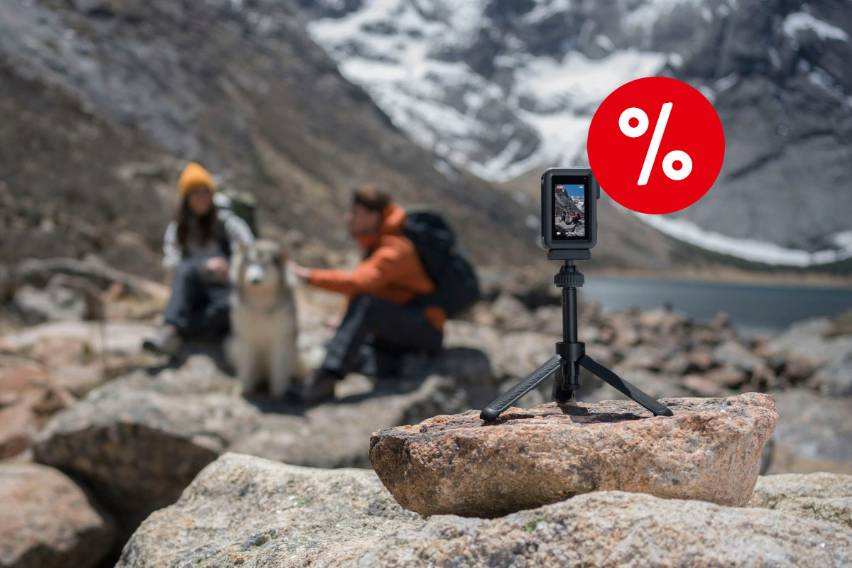 Rechts im Vordergrund eine kleine schwarze hochkant aufgestellte DJI Osmo Action 4 auf kleinem Dreibein-stativ auf Felsen platziert, im Hintergrund zwei Menschen sitzend mit Rucksäcken und weiß grauem Hund vor grau weißer Berglandschaft mit rotem Prozentbutton oben rechts