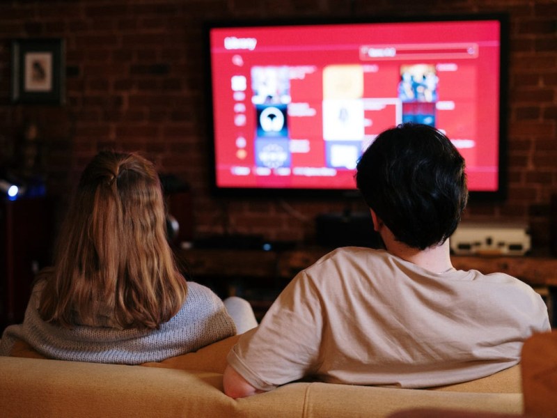 Frau von hinten mit langen hellbraunen Haaren sitzt zusammen mit Mann mit dunklen Haaren auf heller Couch vor Fernseher mit rotem Bildschirm