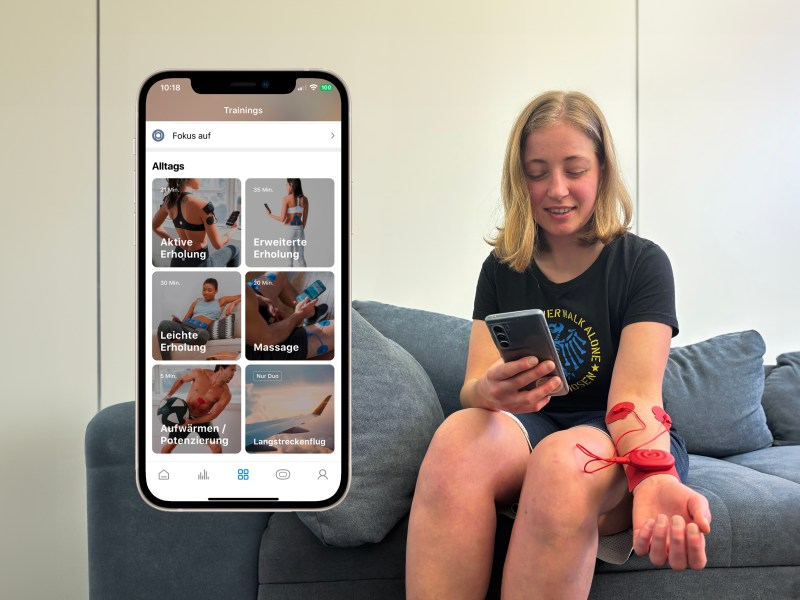 Eine blonde Frau sitzt. mit dem roten Powerdot am Arm auf einem grauen Sofa. Davor ist ein Screenshot der passenden App zu sehen.