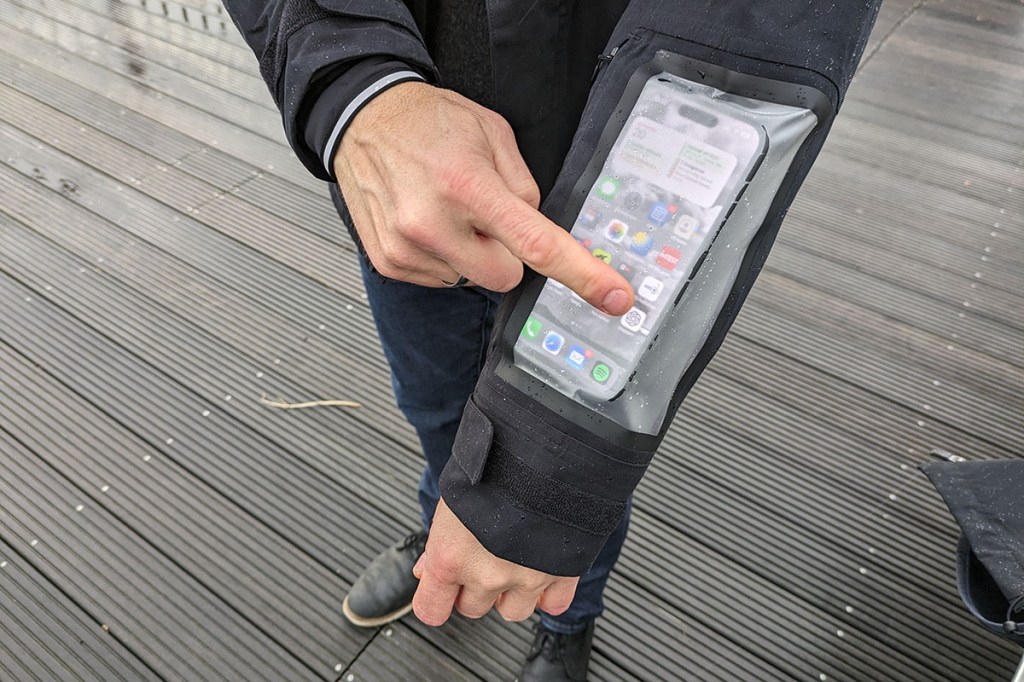 Man trägt Regenjacke, die eine Tasche am Ärmel für ein Smartphone hat. Das eingesteckte Gerät lässt sich durchn eine Klarsichthülle bedienen.