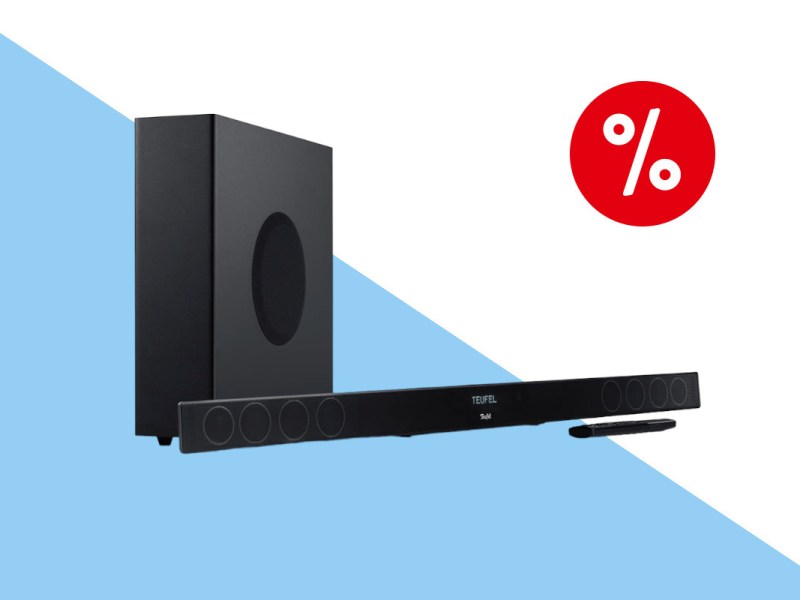 Schwarze längliche Teufel Cinebar 11 Soundbar schräg von vorne, dahinter eine hochkant gestellte schwarze Box auf wei blauem Hintergrund mit rotem Prozentzeichen oben rechts