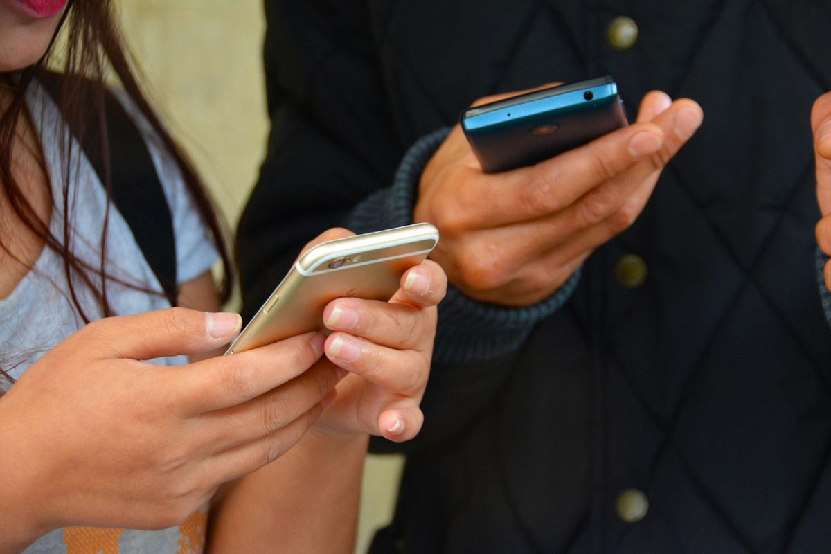 Zwei Paar Hände, links von Frau, rechts von Mann, halten jeweils ein Smartphone (gold, blau), die Frau hat ein hellblaues shirt an, der Mann eine dunkelblaue Jacke