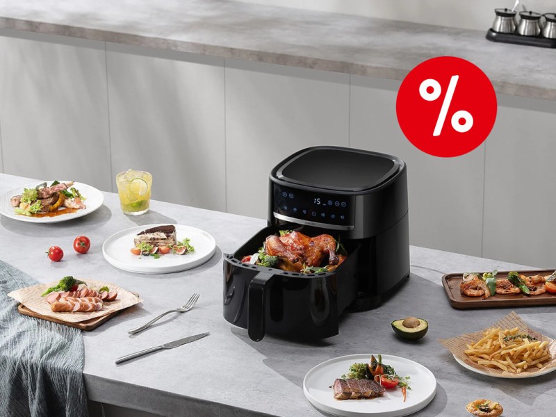 Schwarzer geöffneter Xiaomi Air Fryer mit Hähnchen im Garkorb schräg von oben auf grauer Arbeitsfläche zwischen Tellern mit gegarten Lebensmitteln mit rotem Prozentbutton rechts oben