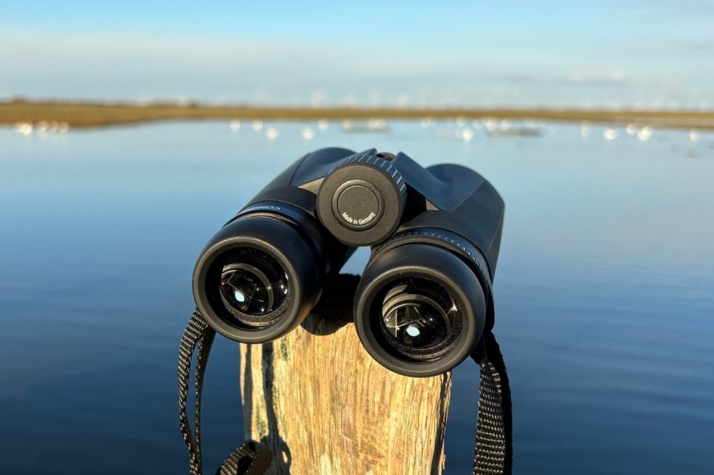 Ein Fernglas von Zeiss, auf einem Holzpfosten liegend, dahinter ein See