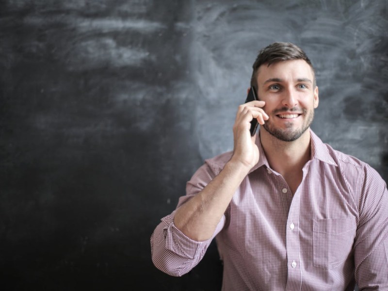 Junger Mann mit Bart und braunen kurzen Haaren lächelnd hält Smartphone ans Ohr in rot weiß kariertem Hemd vor grau schwarzer Wand