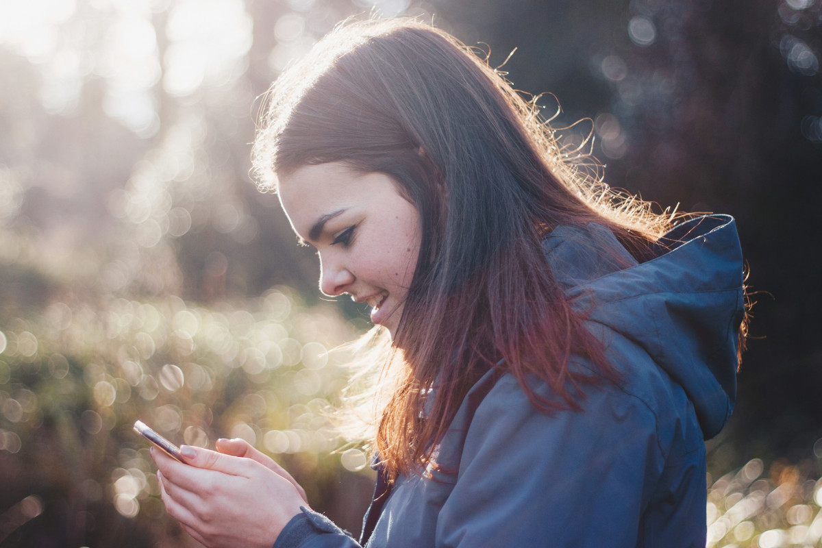 Junge Frau von der Seite lächelnd mit langen braunen Haaren in blauer Softshell-Jacke hält Handy in der Hand, im Hintergrund Sonnenschein und Natur