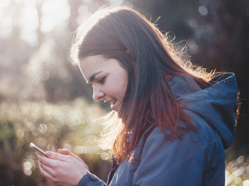 Junge Frau von der Seite lächelnd mit langen braunen Haaren in blauer Softshell-Jacke hält Handy in der Hand, im Hintergrund Sonnenschein und Natur