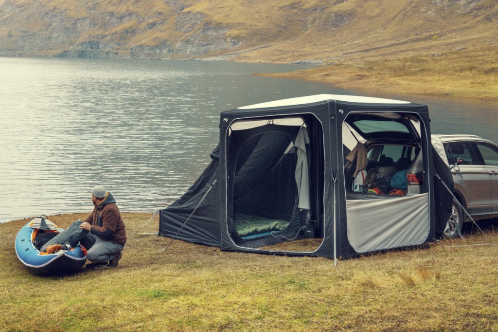 Quadratisches grau weißes Zelt an Heck von silbernem SUV mit geöffneten Türen und Fenstern auf Rasen vor See, Mann, der links vor einem blauem Schlauchboot hockt