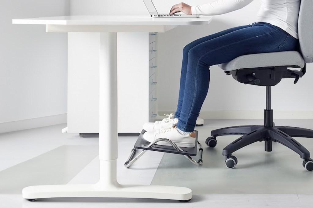 Eine Person sitzt an einem weißen Schreibtisch und benutzt eine IKEA-Fußstütze.