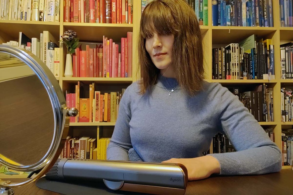 Die Testerin schaut in einen Handspiegel. Die Haare sind komplett geglättet. Hinter ihr ist eine bunte Bücherwand zu sehen.
