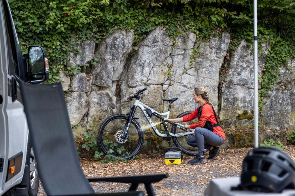 Frau reinigt ihr Fahrrad mit einem Niederdruckreiniger, Fahrrad lehnt vor einer Felswand, im vordergrund sieht man im Anschnitt einen Camper