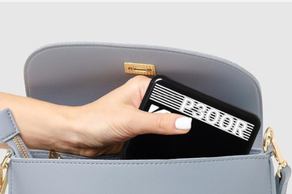 Hand mit weiß lackierten Fingernägeln von links ins Bild hält Schwarzen Mini-Drucker in geöffneter grauer Handtasche
