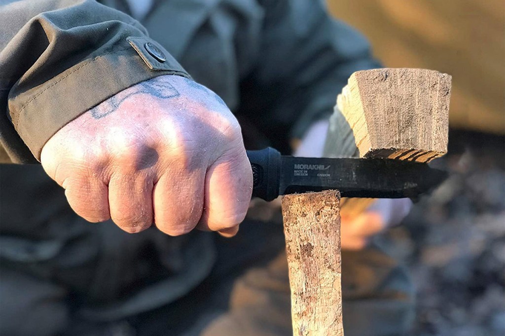 Mann nutzt Outdoormesser zum Spalten eines Holzscheites, in dem er die Messerklinge mit einem Holzstück in den Scheit treibt.