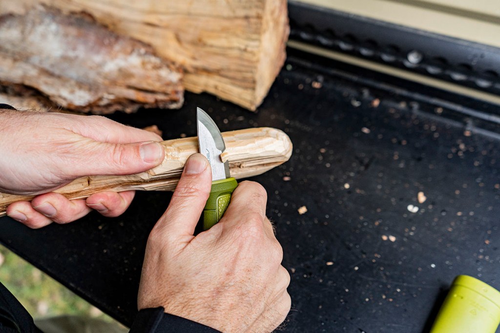 Mann schnitzt mit einem kleinen Outdoormesser in Taschenmessergröße an einem Holzstück.