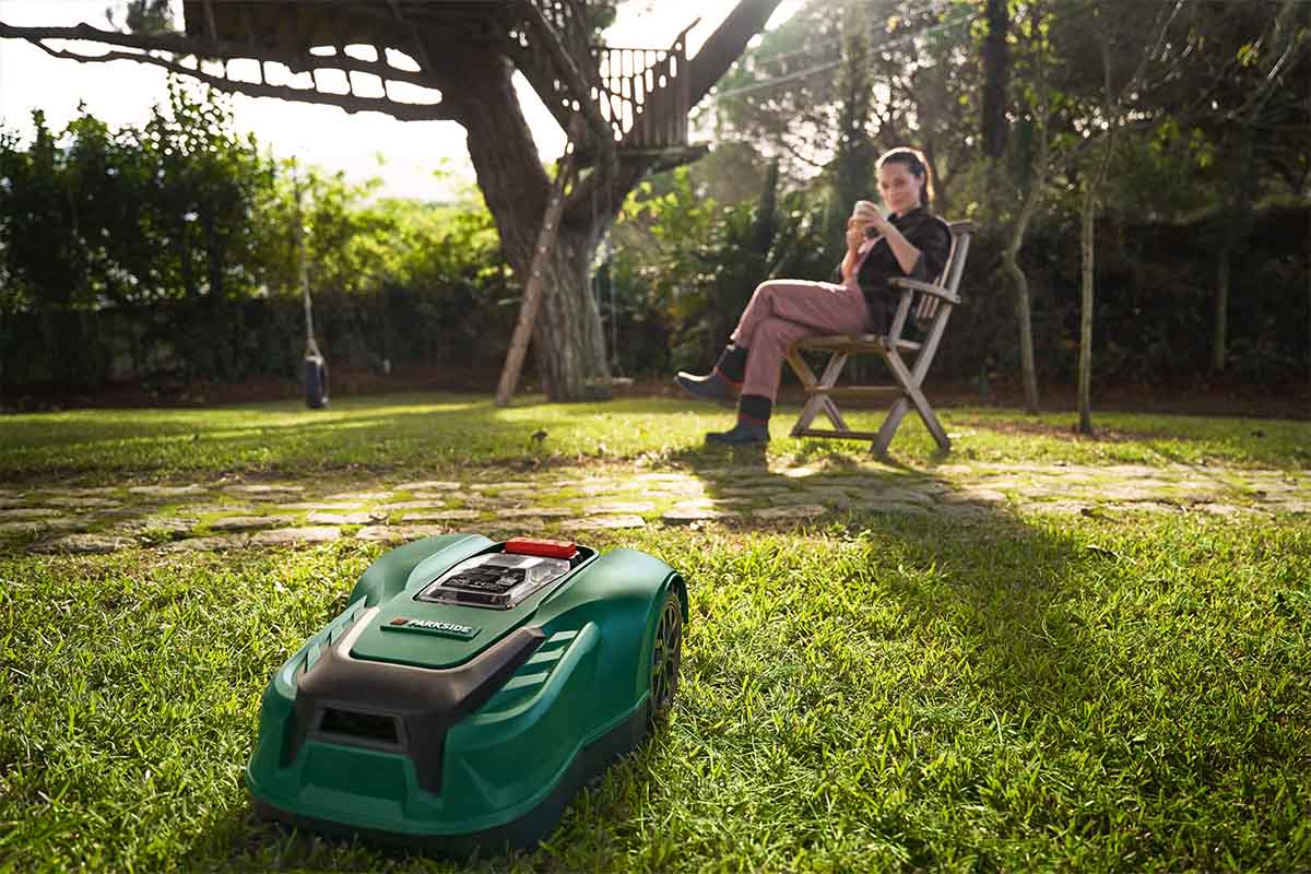 Eine Frau sitzt bequem in einem Stuhl in einem sonnigen Garten und trinkt einen Tee. Im Vordergrund fährt ein Parkside-Mähroboter.