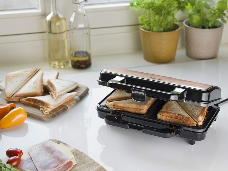 Ein Sandwichmaker auf einer Arbeitsplatte in der Küche. Daneben fertige Sandwiches und Zutaten.