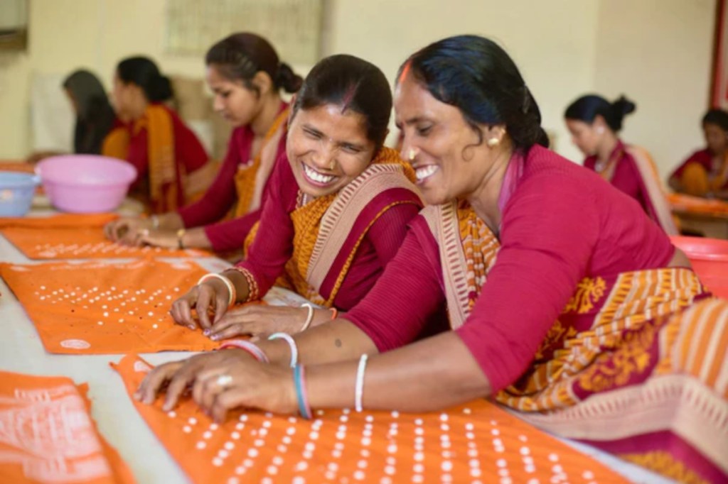 Eine Reihe von rot orange gekleideten indischen Frauen, lachend vor langem Tisch auf dem oranger Stoff liegt