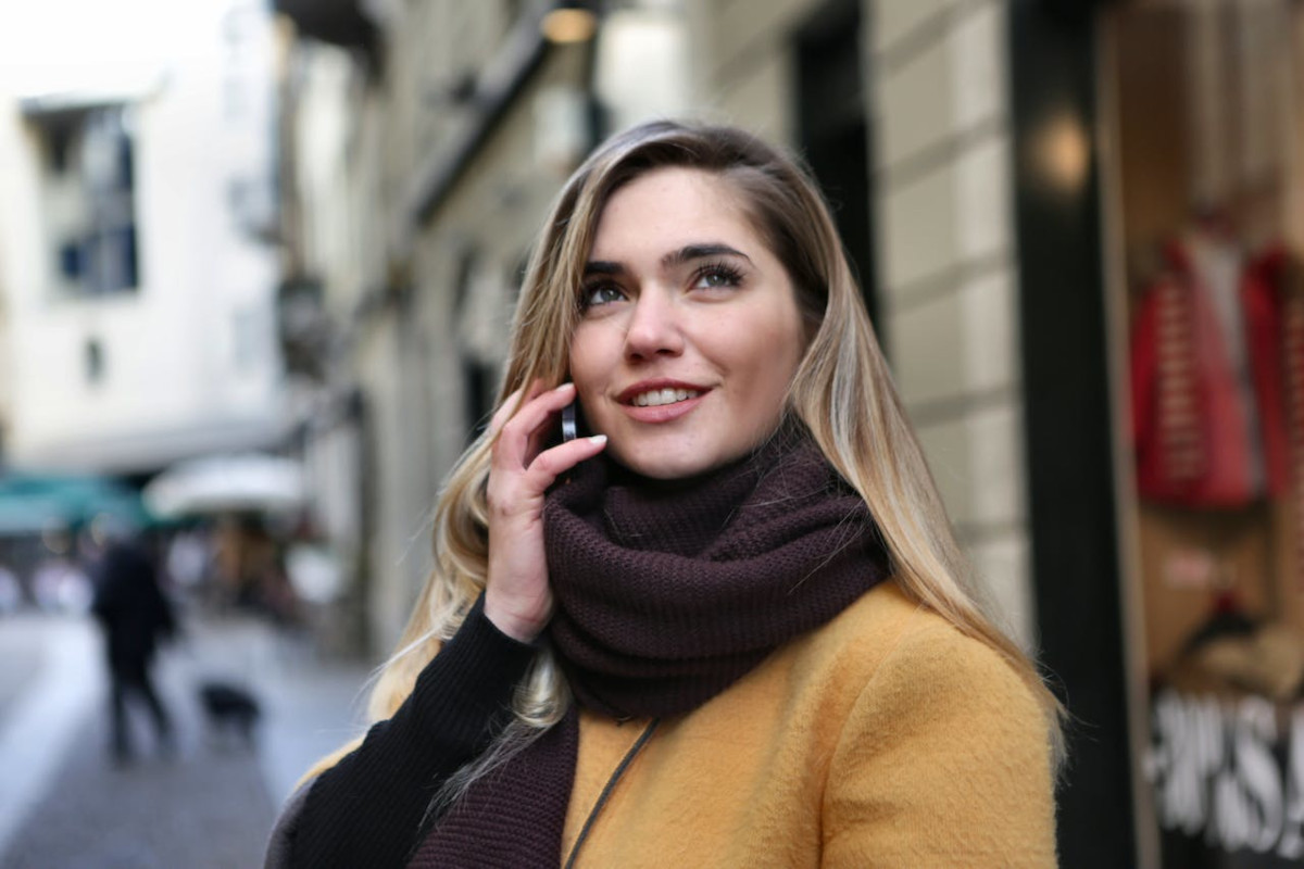 Junge Frau mit blondierten langen Haaren lächelt und hält sich Smartphone ans Ohr in braunem Schal und gelber Stoffjacke vor Altbau