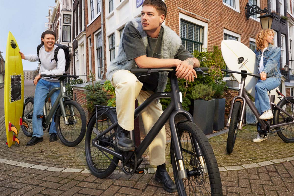 Drei Personen mit ihren E-Bikes, zwei von ihnen haben ein Surfbrett, in einer städtischen Umgebung