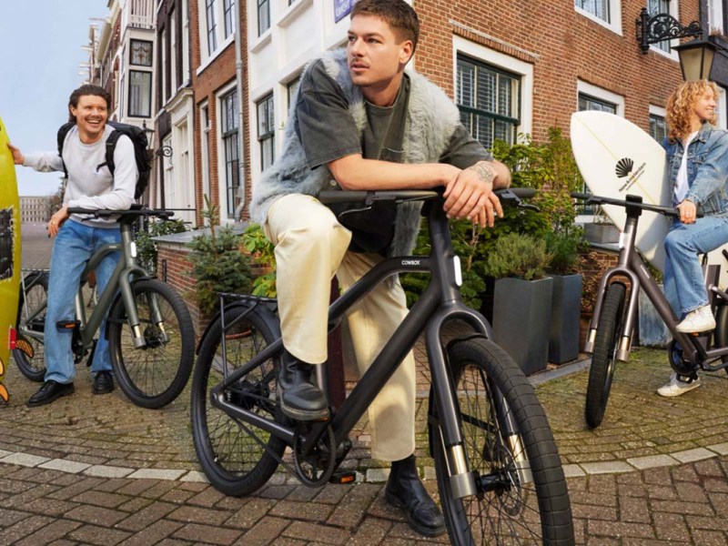 Drei Personen mit ihren E-Bikes, zwei von ihnen haben ein Surfbrett, in einer städtischen Umgebung
