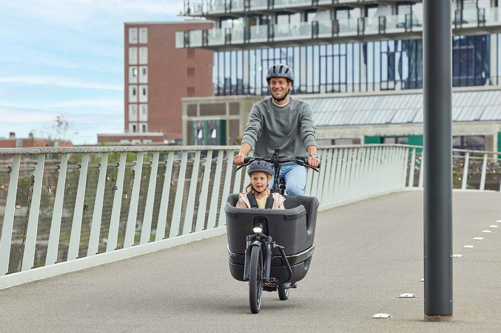 Person fährt mit einem Cargo-E-Bike über eine Brücke, in der Wanne sitzt ein Kind