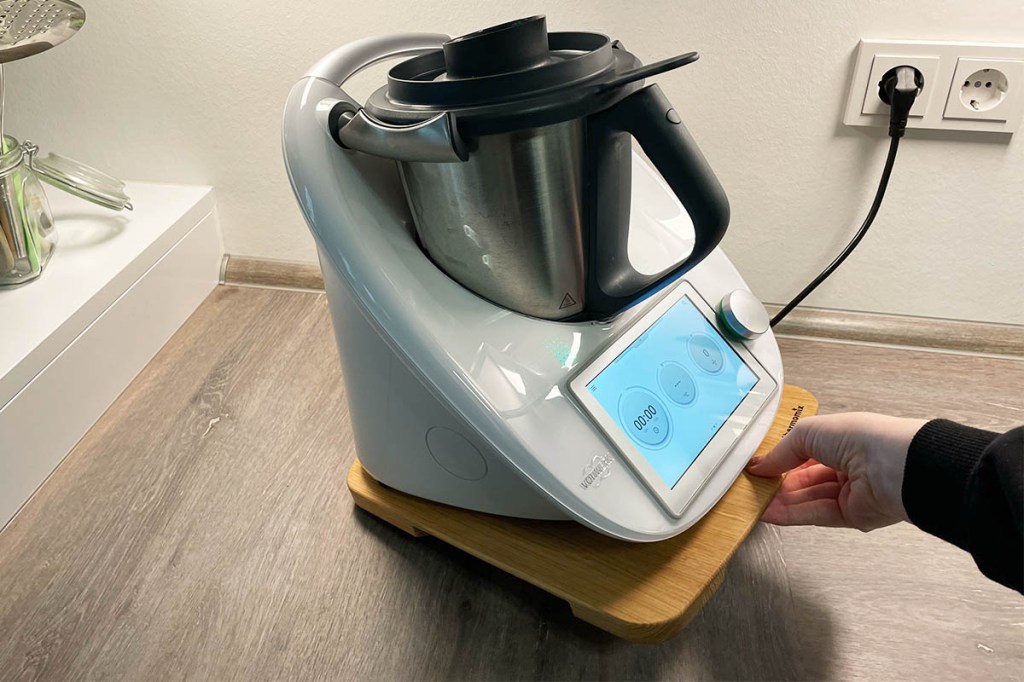 Der Thermomix TM6 mit eingeschaltetem Display auf dem Vorwerk Gleitbrett. Eine Hand hebt das Brett vorne an.