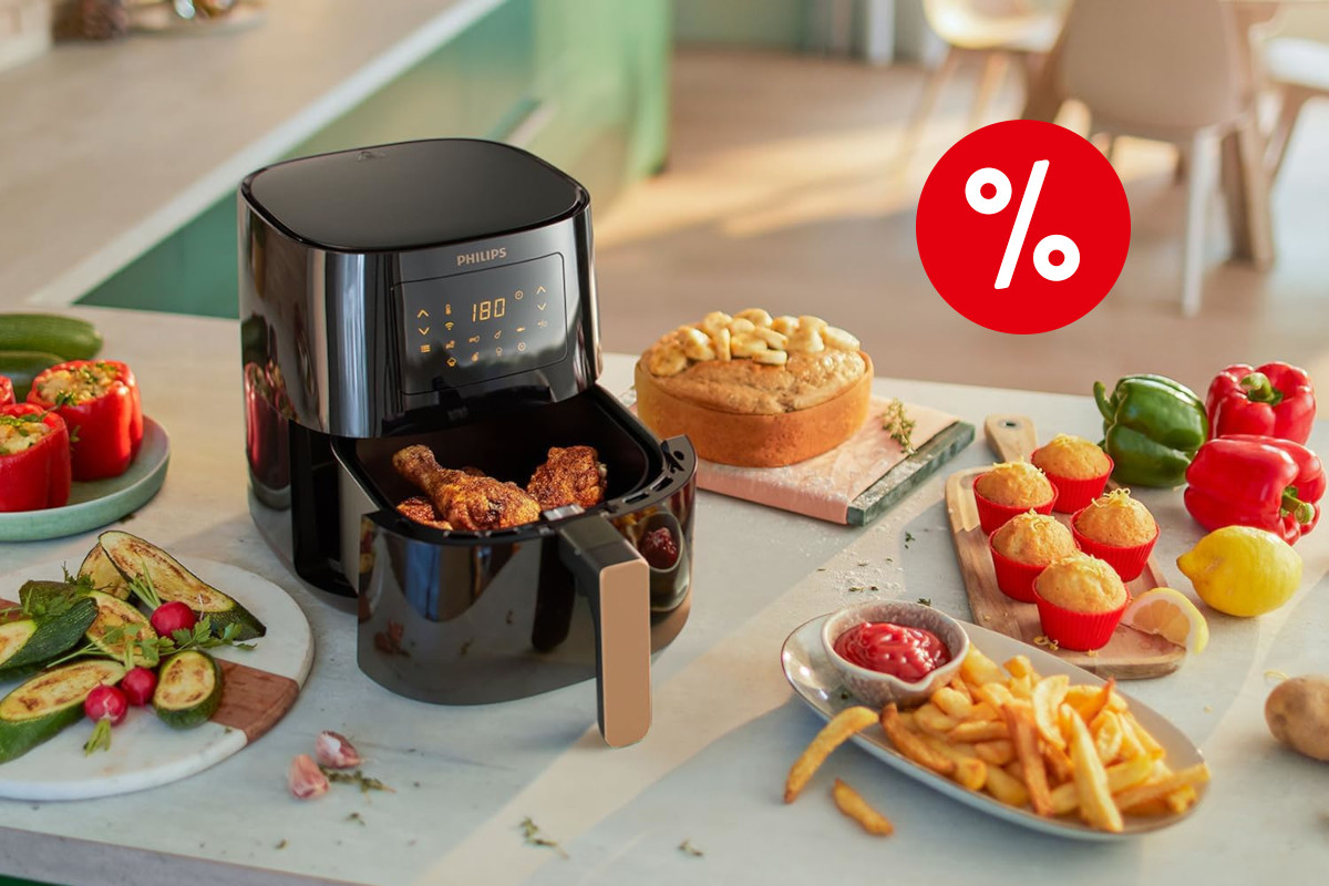 Schwarzer Philips Airfryer mit geöffnetem Garkorb aus dem frittierte Hähnchenbeine ragen auf heller Arbeitsfläche zwischen Tellern mit sämtlichen gegarten Lebensmitteln wie Pommes und Gemüse mit rotem Prozentbutton oben rechts