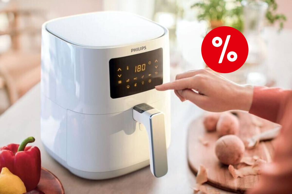 Weißer Philips Airfryer Essential schräg von vorne mit leuchtendem Display auf heller Arbeitsfläche zwischen Paprika und Zwiebeln, von rechts kommt eine Hand rein, um das Gerät zu bedienen, oben rechts ein roter Prozentbutton