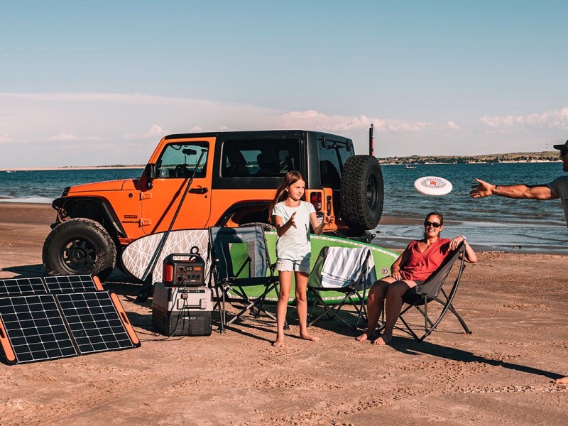 Familie spielt Frisbee am Strand vor orangenem Geländewagen, aufgestellten Solarpanels, Powerstation und Surfbrett