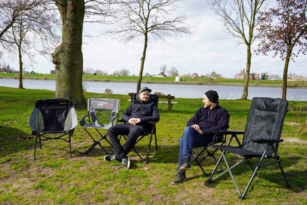 Fünf Campingstühle auf einer Wiese vor einem Fluss. In zwei der Stühle sitzen zwe Männer und unterhalten sich.