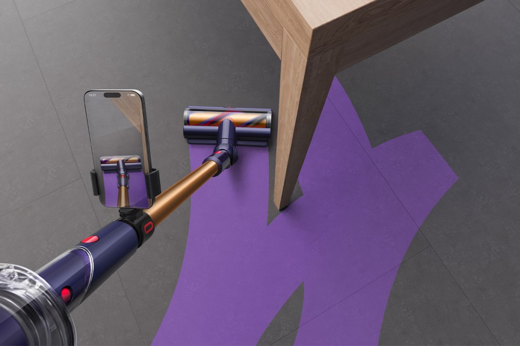 Ein Dyson-Akkusauger saugt um einen Tisch herum. Ein Smartphone ist am Saugstiel angebracht. Es zeigt lila Streifen, die auch auf dem Boden eingezeichnet sind.
