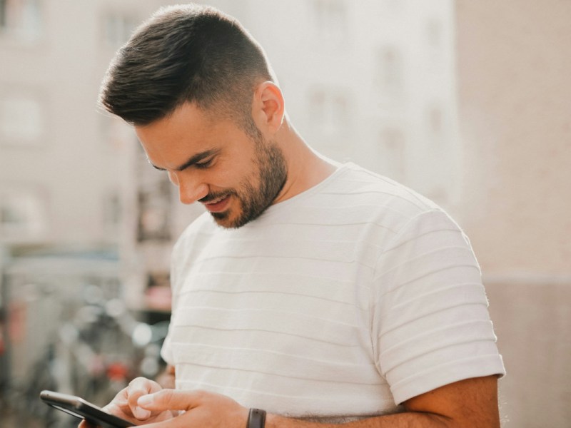 Lächelnder Mann mit dunklen kurzen Haaren und Bart in weißem Tshirt schaut auf Smartphone, vor heller freundlicher Stadtkulisse