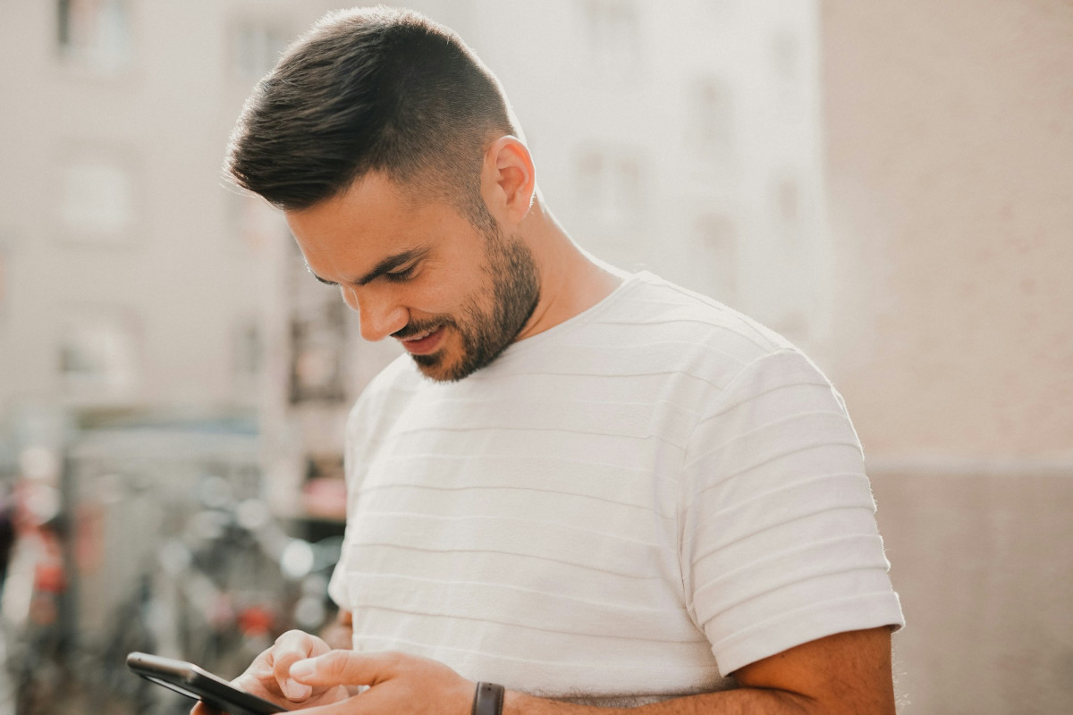 Lächelnder Mann mit dunklen kurzen Haaren und Bart in weißem Tshirt schaut auf Smartphone, vor heller freundlicher Stadtkulisse