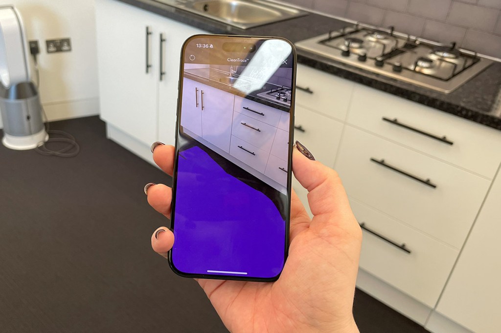 Eine Hand hält ein Smartphone vor die Kamera. Darauf ist das im Hintergrund befindliche Küchenszenario zu sehen. Der Boden ist auf dem Handy allerdings überwiegend lila statt grau.