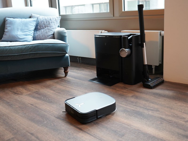 Der Ecovacs Deebot X2 Saugwischroboter in Wohnzimmer-Umgebung. Im Hintergrund steht die Combo-Station mit Handstaubsauger und Akkusauger-Aufsatz.