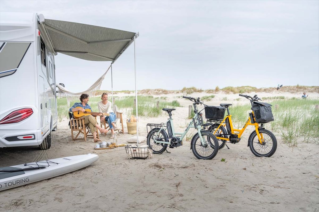 Anschnitt Camper an einem Strand stehend, zwei Menschen sitzen auf Stühlen davor, zwei Kompakt-E-Bikes im Vordergrund