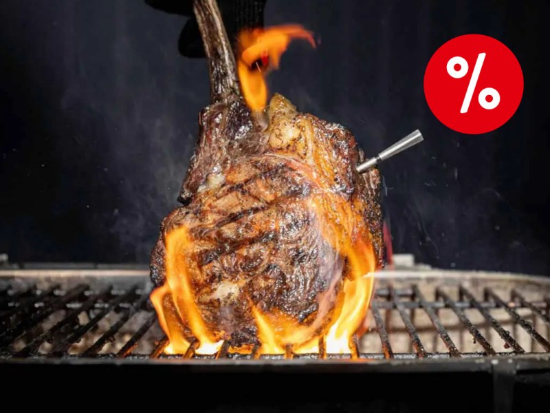Gegrilltes Stück Fleisch wird am Knochen senkrecht gehalten auf Grillrost mit Flammen ringsum und silberner dünne längliche Spitze von Meater 2 Plus Thermometer rausragend vor dunklem Hintergrund mit rotem Prozentbutton oben rechts