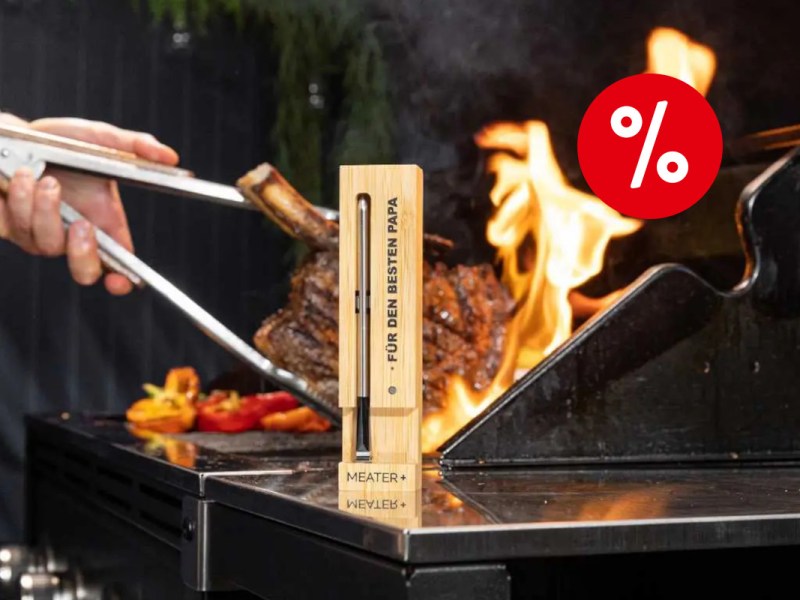 In der Mitte eine schmale senkrecht hingestellte Holzbox mit schmalem silbernem Fleischthermometer Meaer Plus drin, am Rand aufgedruckt "Für den besten Papa" auf silberner Arbeitsfläche, dahinter Grill in dem ein Fleischstück von silberner Zange umringt von Flammen gewendet wird mit rotem Prozentbutton oben rechts