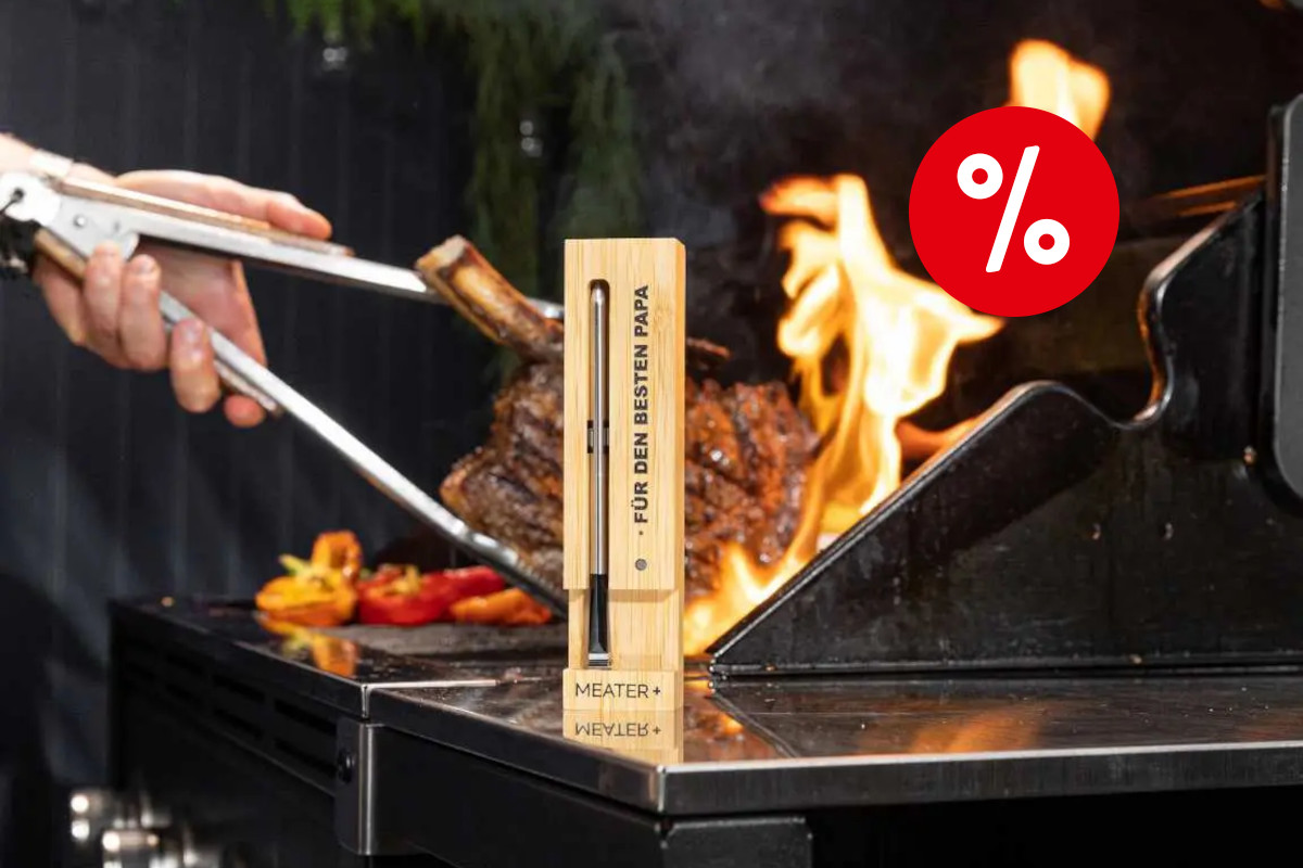 In der Mitte eine schmale senkrecht hingestellte Holzbox mit schmalem silbernem Fleischthermometer Meaer Plus drin, am Rand aufgedruckt "Für den besten Papa" auf silberner Arbeitsfläche, dahinter Grill in dem ein Fleischstück von silberner Zange umringt von Flammen gewendet wird mit rotem Prozentbutton oben rechts