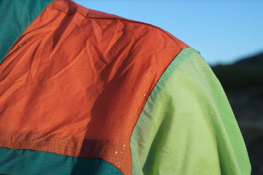 Detail von Schulter mit neonbunter Jacke auf der Wassertropfen abperlen vor blauem Himmel und dunklen Bergen