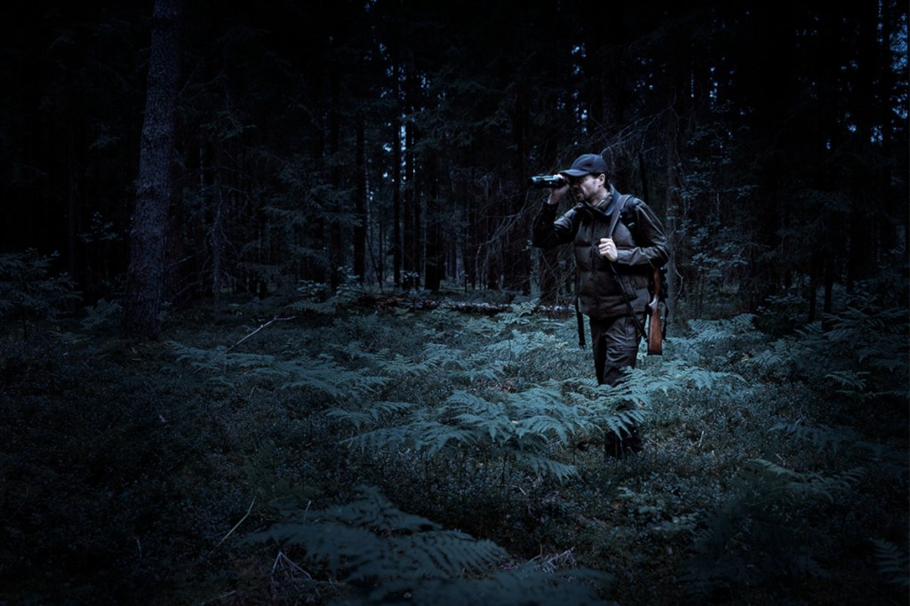 Dunkles Bild von Waldlichtung mit dunkelangezogenem Mann mit Gewehr über Schulter, der durch ein schwarzes längliches Nachtsichtgerät schaut