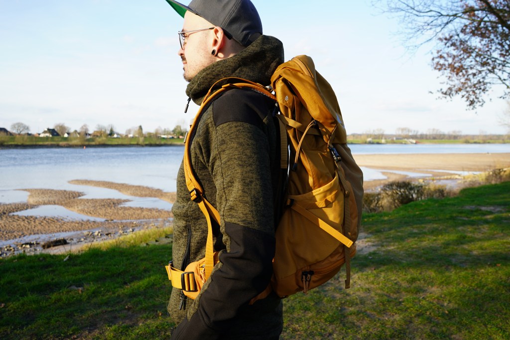 Mann steht seitlich zum Fotografen, er hat einen gelben Rucksack auf, im Hintergrund ein Fluss