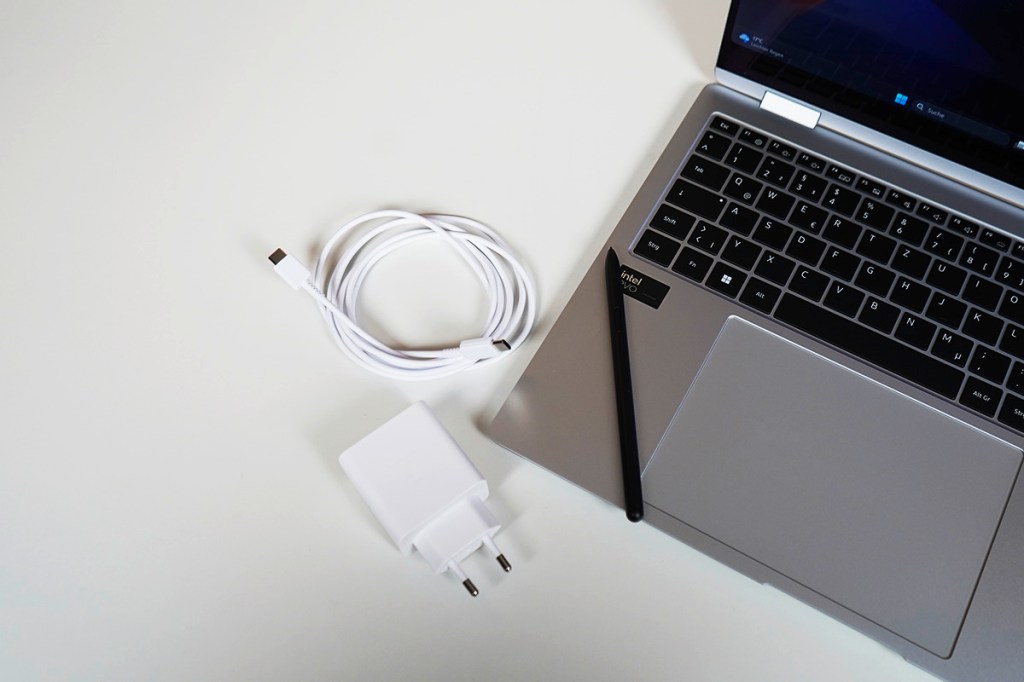 Auf einem weißen Tisch liegt ein weißes Netzteil, ein weißes Kabel und ein schwarzer Stift neben einem aufgeklappten Notebook.