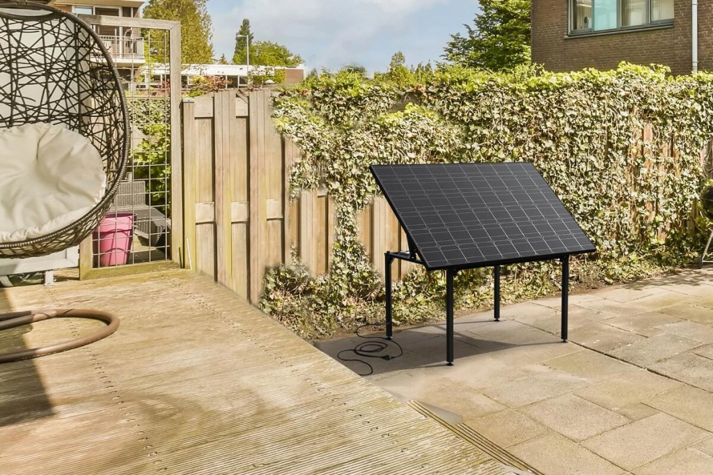 Schwarzes Solarmodul aufgestellt auf vierbeinigem Tischgestell auf weiter Terrasse mit hellem Steinboden vor hellem Holzzaun an dem Rankenpflanze wächst