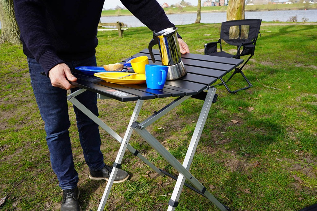 Mann hebt Campingtisch auf Campingplatz an, dabei knickt der Tisch in der Mi9tte zusammen. Teller und Tassen verrutschen.